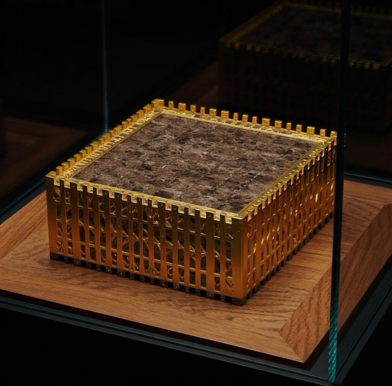K&ouml;nnte beinhalten: Eine goldfarbene, rechteckige Box mit braunem Innenraum, ausgestellt auf einem Holzsockel. Die Box hat ein dekoratives zaun&auml;hnliches Design an den Seiten und ist in einer Glasvitrine eingeschlossen.