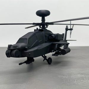 May include: A black model helicopter with detailed features, including rotors, a cockpit, and weaponry. The miniature aircraft is positioned on a gray surface, showcasing its design and structure. The model is a detailed replica of a military helicopter.