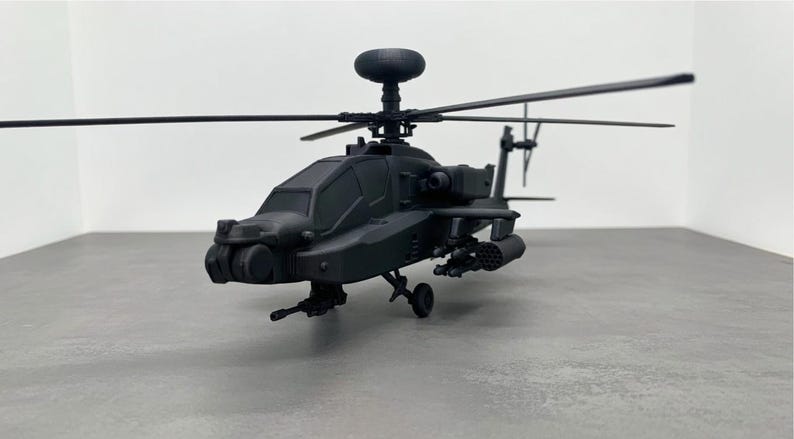 May include: A black model helicopter with detailed features, including rotors, a cockpit, and weaponry. The miniature aircraft is positioned on a gray surface, showcasing its design and structure. The model is a detailed replica of a military helicopter.