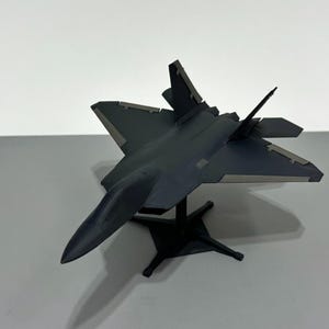 May include: A dark gray model airplane, possibly a fighter jet, is displayed on a black stand. The aircraft has angled wings and a sleek design. The model is set against a plain white background.