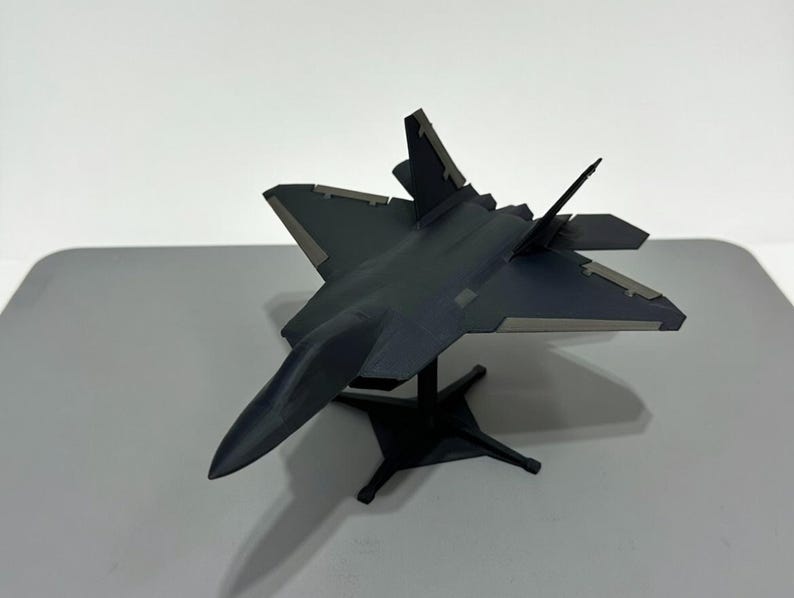 May include: A dark gray model airplane, possibly a fighter jet, is displayed on a black stand. The aircraft has angled wings and a sleek design. The model is set against a plain white background.