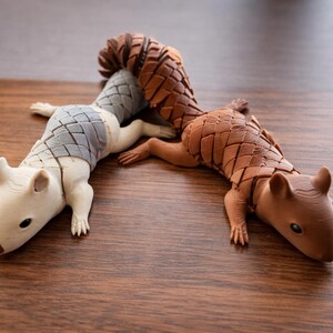 May include: Two decorative pangolin figurines. One is off-white with a gray segmented back, the other is brown with a brown segmented back. Both are lying on a wooden surface.