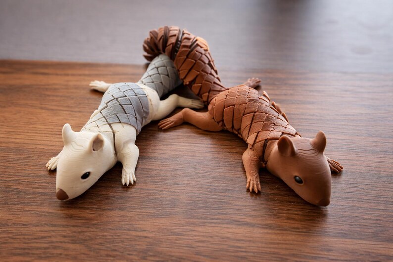 May include: Two decorative pangolin figurines. One is off-white with a gray segmented back, the other is brown with a brown segmented back. Both are lying on a wooden surface.