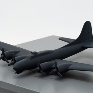 May include: A matte black model airplane with four propellers and a tall tail fin. The model is a replica of a vintage bomber aircraft, resting on a gray surface. A small black rectangular block sits to the left of the plane.