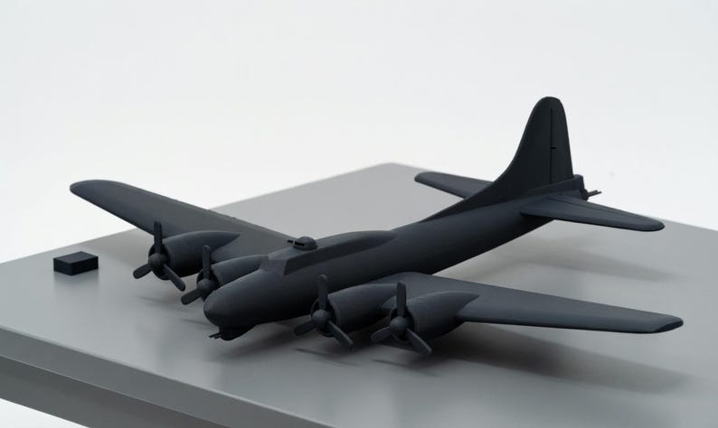 May include: A matte black model airplane with four propellers and a tall tail fin. The model is a replica of a vintage bomber aircraft, resting on a gray surface. A small black rectangular block sits to the left of the plane.