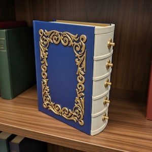 May include: A blue and gold decorative book-shaped box with a gold filigree design on the front. The box has a white spine with four small gold knobs. The box is on a wooden shelf with other books.