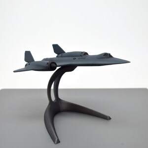 May include: A gray model airplane, a SR-71 Blackbird, is displayed on a gray stand. The aircraft has a sleek design with wings and a cockpit. The stand has a curved design with three legs. The model is a detailed replica of the iconic aircraft.