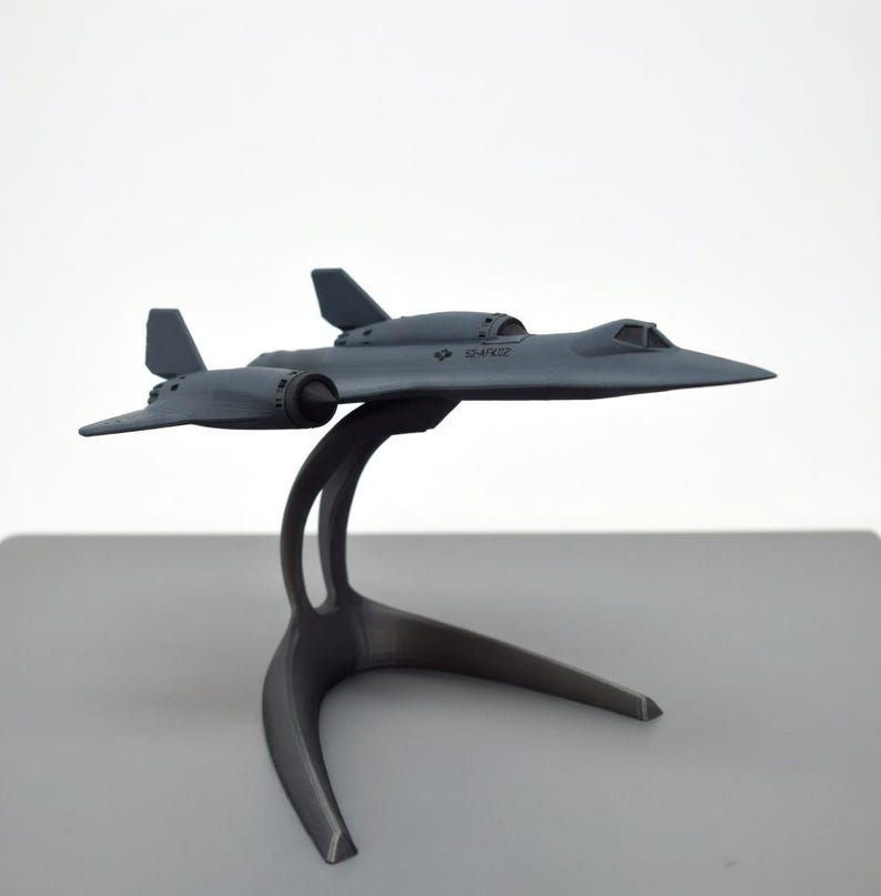May include: A gray model airplane, a SR-71 Blackbird, is displayed on a gray stand. The aircraft has a sleek design with wings and a cockpit. The stand has a curved design with three legs. The model is a detailed replica of the iconic aircraft.