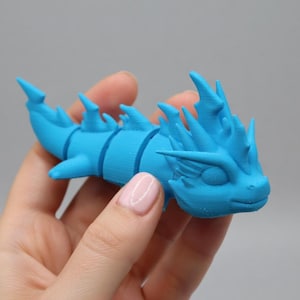 May include: A light blue, articulated dragon figurine. The dragon has a segmented body, fins, and a detailed head with spikes. The figurine is held in a hand against a gray background. The dragon is a toy or decorative item.