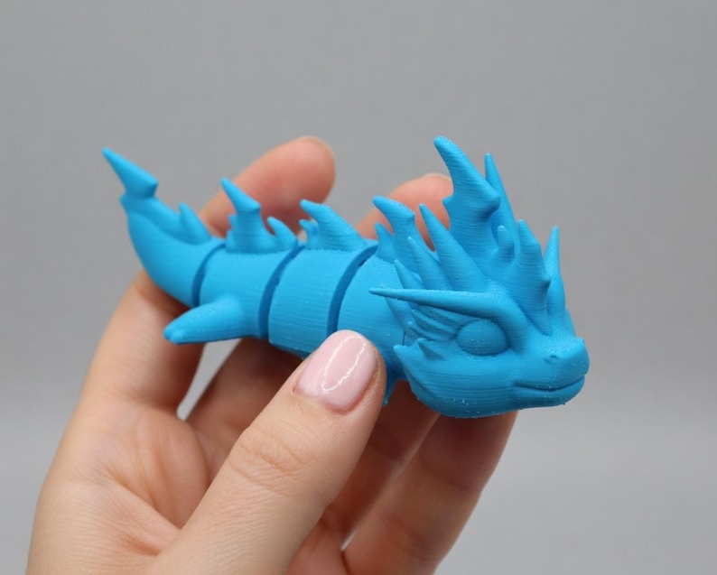 May include: A light blue, articulated dragon figurine. The dragon has a segmented body, fins, and a detailed head with spikes. The figurine is held in a hand against a gray background. The dragon is a toy or decorative item.