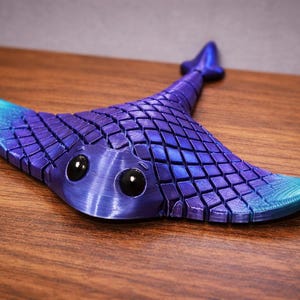 May include: A 3D-printed manta ray figurine in shades of blue and purple. The ray has a textured, tiled pattern on its body, with large, black eyes. The tips of the wings are a lighter blue. The figurine is on a wooden surface.