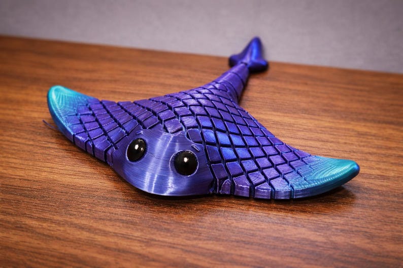 May include: A 3D-printed manta ray figurine in shades of blue and purple. The ray has a textured, tiled pattern on its body, with large, black eyes. The tips of the wings are a lighter blue. The figurine is on a wooden surface.