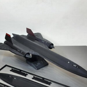 May include: A black SR-71 model airplane with red accents on the tail fins. The model is displayed on a stand with the text "SR-71". Additional unassembled parts are visible in the foreground. The model is a detailed replica.