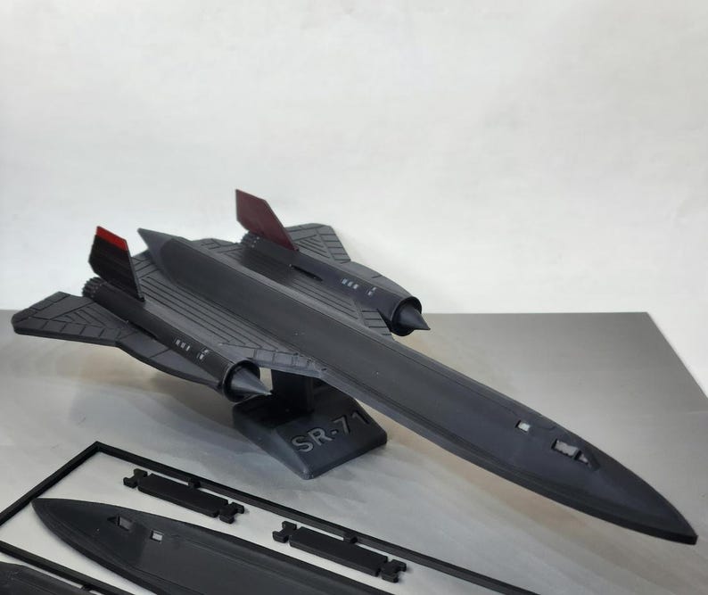 May include: A black SR-71 model airplane with red accents on the tail fins. The model is displayed on a stand with the text "SR-71". Additional unassembled parts are visible in the foreground. The model is a detailed replica.