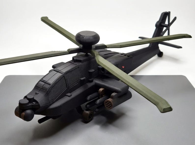 May include: A detailed model of a black helicopter with olive green rotor blades. The model features intricate detailing, including the cockpit, weapons, and tail rotor. The helicopter is positioned on a gray surface.