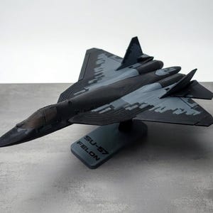 May include: A detailed model of an SU-57 FELON fighter jet, predominantly black and gray with camouflage patterns on the wings and fuselage. The model sits on a gray base with the text "SU-57 FELON".