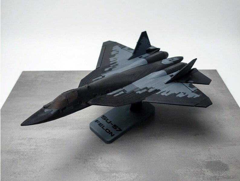 May include: A detailed model of an SU-57 FELON fighter jet, predominantly black and gray with camouflage patterns on the wings and fuselage. The model sits on a gray base with the text "SU-57 FELON".