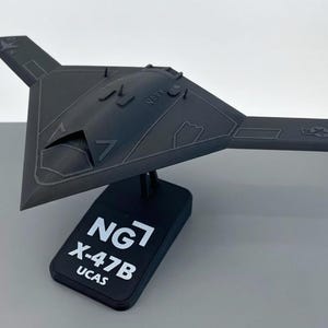 May include: A dark gray model of an X-47B UCAS unmanned combat air system aircraft. The model is mounted on a black base with the text "NG7 X-47B UCAS". The aircraft has a triangular wing shape and the word "NAVY" is visible.