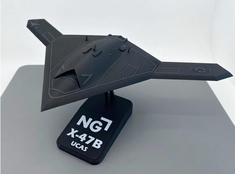 May include: A dark gray model of an X-47B UCAS unmanned combat air system aircraft. The model is mounted on a black base with the text "NG7 X-47B UCAS". The aircraft has a triangular wing shape and the word "NAVY" is visible.