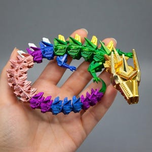 May include: A colorful articulated dragon toy in shades of pink, purple, blue, green, and gold. The dragon is held in a hand, showcasing its flexible design. The head is gold, and the body transitions through a rainbow of colors.