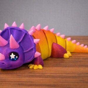 May include: A colorful 3D-printed triceratops figurine. The dinosaur features a purple head with pink horns and spikes, transitioning to a yellow and orange body. It has large eyes with star-shaped pupils and is lying down.