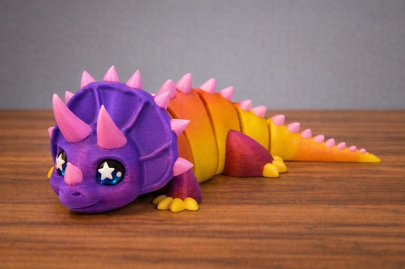 May include: A colorful 3D-printed triceratops figurine. The dinosaur features a purple head with pink horns and spikes, transitioning to a yellow and orange body. It has large eyes with star-shaped pupils and is lying down.