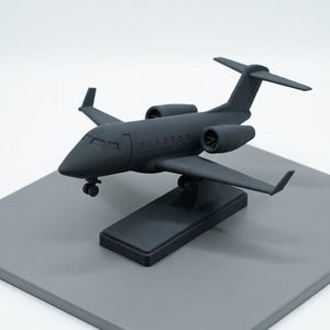 May include: A matte black model airplane, a miniature replica of a jet, is displayed on a gray rectangular base. The model features detailed wings, engines, and a cockpit, showcasing a sleek, modern design. The airplane is a decorative item.
