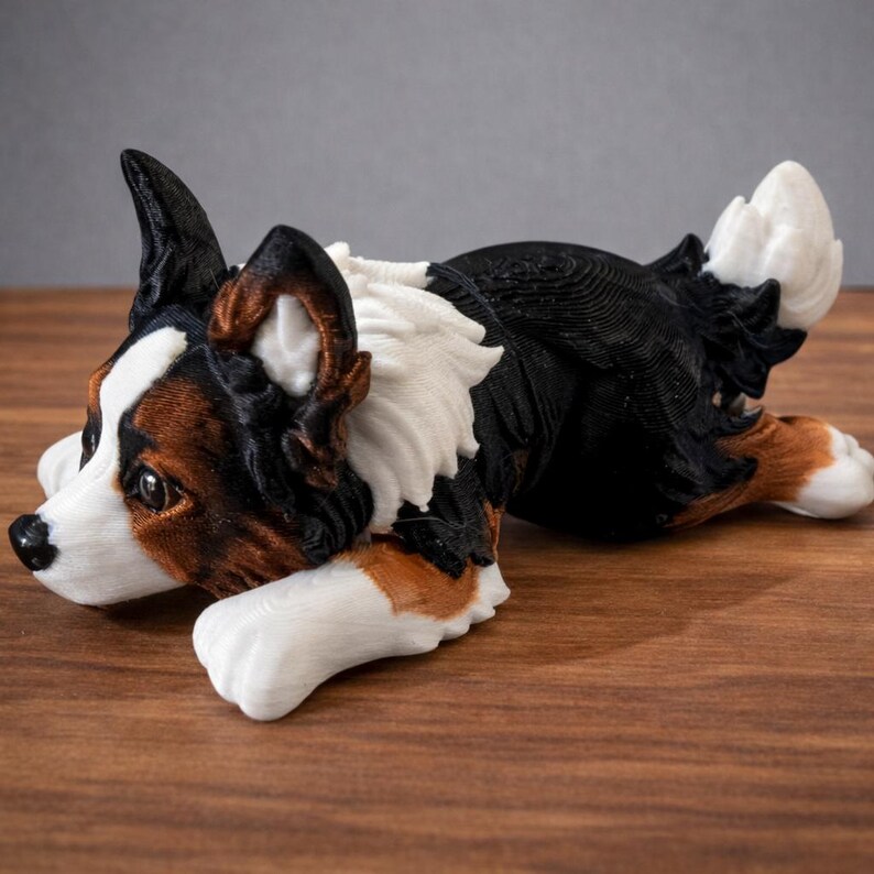 May include: A 3D-printed dog figurine in a relaxed pose. The dog has a white, black, and brown coat, with detailed fur textures. The figurine is lying on a wooden surface, showcasing its realistic design. A decorative item for dog lovers.