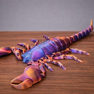 May include: A detailed, iridescent scorpion sculpture in shades of purple, blue, and gold. The arthropod has a segmented body, pincers, and a curved tail. The sculpture is displayed on a wooden surface.