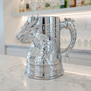 A professional collage showcasing over 20 unique 3D printable STL models for beer mugs and can holders. The image features high-detail designs including a horned Minotaur with gold accents, a gothic skull wearing aviator goggles, and a steampunk gas mask style tankard with copper pipes. The background displays various mythical, viking, and dark fantasy themed mugs in metallic silver and bronze finishes. Large yellow and blue 3D text in the center reads '20+ Can Holder & Beer Mug STL'.