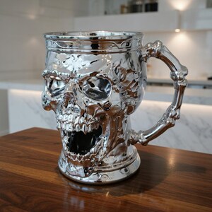 A professional collage showcasing over 20 unique 3D printable STL models for beer mugs and can holders. The image features high-detail designs including a horned Minotaur with gold accents, a gothic skull wearing aviator goggles, and a steampunk gas mask style tankard with copper pipes. The background displays various mythical, viking, and dark fantasy themed mugs in metallic silver and bronze finishes. Large yellow and blue 3D text in the center reads '20+ Can Holder & Beer Mug STL'.