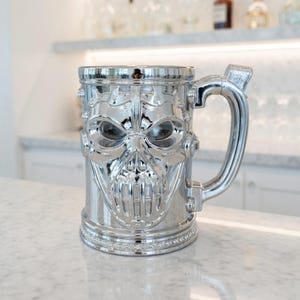 A professional collage showcasing over 20 unique 3D printable STL models for beer mugs and can holders. The image features high-detail designs including a horned Minotaur with gold accents, a gothic skull wearing aviator goggles, and a steampunk gas mask style tankard with copper pipes. The background displays various mythical, viking, and dark fantasy themed mugs in metallic silver and bronze finishes. Large yellow and blue 3D text in the center reads '20+ Can Holder & Beer Mug STL'.