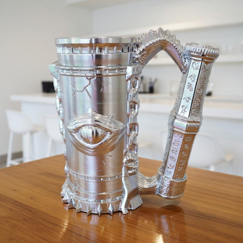 A professional collage showcasing over 20 unique 3D printable STL models for beer mugs and can holders. The image features high-detail designs including a horned Minotaur with gold accents, a gothic skull wearing aviator goggles, and a steampunk gas mask style tankard with copper pipes. The background displays various mythical, viking, and dark fantasy themed mugs in metallic silver and bronze finishes. Large yellow and blue 3D text in the center reads '20+ Can Holder & Beer Mug STL'.