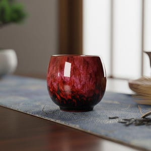 Op de afbeelding: Een kleine, ronde, rood-zwarte glazen theekop met een textuur. De kop staat op een blauw patroon tafelloper. De theekop heeft een glanzende afwerking en is ontworpen om thee te drinken.