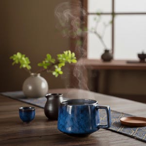 Könnte beinhalten: Eine blaue Keramiktasse mit Henkel, gefüllt mit dampfender Flüssigkeit, steht auf einem Holztisch. Eine kleine blaue Tasse, ein dunkelbrauner Krug und eine kleine Vase mit grünen Zweigen stehen ebenfalls auf dem Tisch. Ein Holztablett und ein gemusterter Tischläufer vervollständigen die Szene.