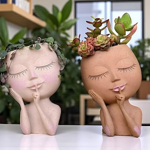 Puede incluir: Dos macetas de cerámica con forma de rostro, una rosa claro y otra marrón claro, cada una con los ojos cerrados y un dedo en los labios. La maceta rosa claro tiene vegetación, y la marrón claro tiene suculentas.