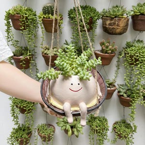 Puede incluir: Una maceta de cerámica rosa claro con una cara sonriente y piernas colgantes, suspendida por una cuerda. La maceta está llena de suculentas verdes. Otras macetas colgantes de terracota con suculentas están en el fondo.