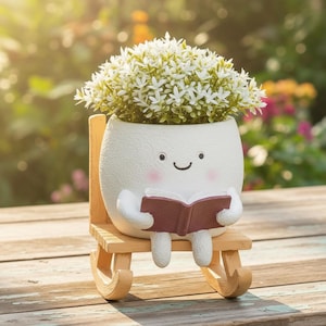 Op de afbeelding: Een grillige witte keramische plantenbak in de vorm van een lachend karakter, met een boek in de hand en zittend op een houten schommelstoel. De plantenbak is gevuld met kleine witte bloemen. De achtergrond is een vage tuin.