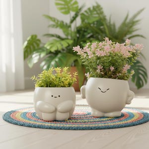 Peut inclure: Deux pots de fleurs en céramique blanche avec des visages de dessins animés, remplis de verdure et de fleurs. L'un est petit et rond, l'autre a une forme humanoïde avec des bras. Ils reposent sur un tapis circulaire coloré, avec de grandes plantes en arrière-plan.