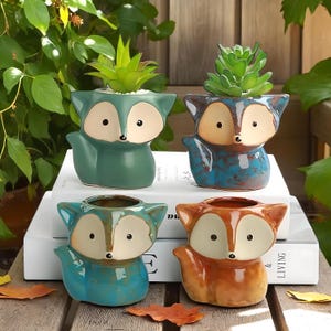 May include: Four ceramic fox-shaped planters in teal, blue, and orange, each with a small succulent. The planters have white faces and black eyes, sitting on a stack of white books.