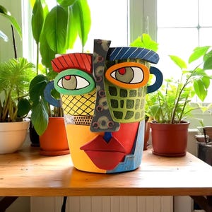 Pode incluir: Um vaso colorido em forma de rosto abstrato com boca vermelha, olhos verdes e laranja e um rosto com padrões amarelos e verdes. O vaso tem alças azuis e está sobre uma mesa de madeira, com plantas ao fundo.