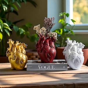 Puede incluir: Tres jarrones en forma de corazón en oro, rojo y blanco, cada uno con flores secas. Los jarrones están colocados sobre una superficie de madera con un ejemplar de la revista Vogue. Los jarrones son de diseño anatómico.
