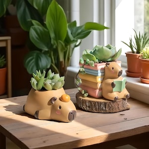 May include: Two novelty capybara-shaped plant pots, each filled with succulents. One is lying down with an orange fruit on its head. The other is sitting and reading a book, with a stack of colourful books behind it. The pots are on a wooden surface.