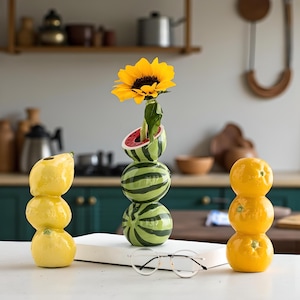 Può includere: Natura morta con tre vasi in ceramica a forma di frutta. Un vaso è composto da limoni impilati, un altro da arance impilate e il terzo da angurie impilate con un girasole. I vasi sono su una superficie bianca con occhiali.