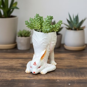 May include: A white fox-shaped planter with a green succulent plant. The fox has red eyes and markings, with orange accents on the ears and paws. The planter is on a wooden surface, with other potted plants in the background.