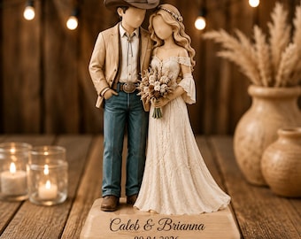 May include: A wedding cake topper figurine featuring a couple standing on a wooden base engraved with the text Caleb & Brianna and 09.04.2026. The figure wears a brown cowboy hat, tan blazer, and blue jeans, while the other wears a cream dress.