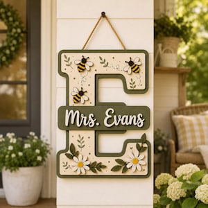 May include: A wooden letter E door sign featuring a cream-colored base with dark green borders. The sign is decorated with painted bees, green leaves, and white daisy flowers. A horizontal dark green bar displays the white text Mrs. Evans.