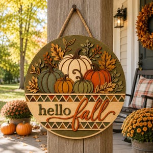 May include: A circular wooden door sign featuring a layered design of pumpkins in shades of cream, orange, and green, surrounded by autumn leaves. The sign includes the text hello fall in dark green and orange script, suspended by a rope.