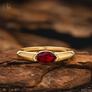 Puede incluir: Un anillo de oro con una piedra preciosa de color rojo intenso en talla marquesa, engastada horizontalmente en el centro. El aro es liso y pulido, estrechándose ligeramente hacia el engaste. El diseño es limpio y destaca el contraste del metal.