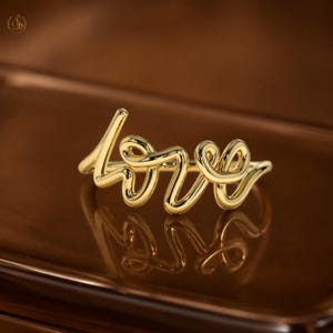 May include: A gold-coloured ring spelling out the word "love" in a cursive font. The ring is set on a brown surface, with its shape reflected. This is a piece of jewellery.